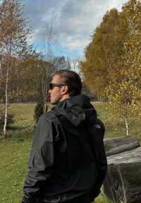 Black waterproof jacket with a hood, featuring a logo on the shoulder. The background includes green grass and autumnal trees.