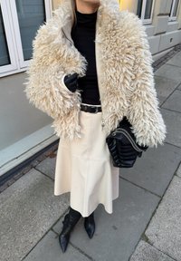 Veste en fourrure shaggy crème, col roulé noir ajusté, jupe en cuir crème, gants noirs et sac matelassé, assortis à des bottines noires à bout pointu.