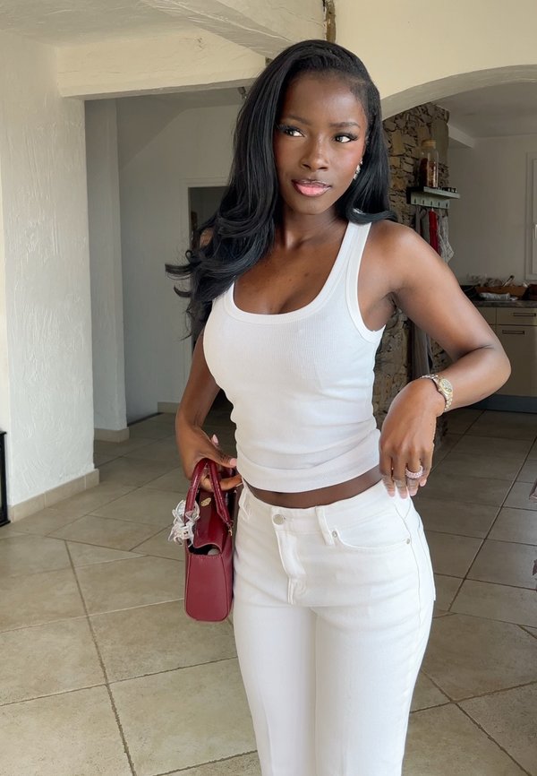 Femme aux longs cheveux noirs portant un débardeur blanc et un pantalon blanc, tenant un sac à main rouge dans une pièce carrelée avec des murs en pierre et blancs.