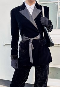 Personne portant un costume en velours noir avec des revers en satin et une ceinture à la taille, des gants assortis avec des poignets en fourrure, tenant un sac à main noir avec une bandoulière blanche.