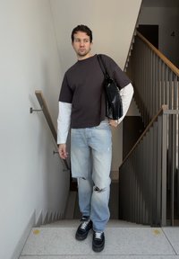 Homme debout sur un escalier portant une chemise à manches longues noire et blanche superposée, un jean clair déchiré, des baskets noires, et portant un sac à bandoulière noir.