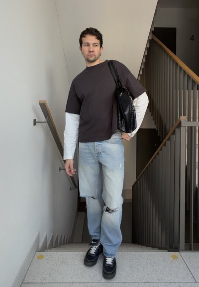 Hombre de pie en una escalera con una camiseta de manga larga en capas blanca y negra, jeans claros rasgados, zapatillas negras y llevando un bolso negro de hombro.