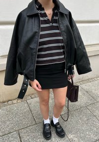 Czarna skórzana kurtka na czarno-białej, paskowanej koszulce polo, w połączeniu z czarną spódnicą, białymi skarpetkami i czarnymi mokasynami, trzymająca brązową torbę.