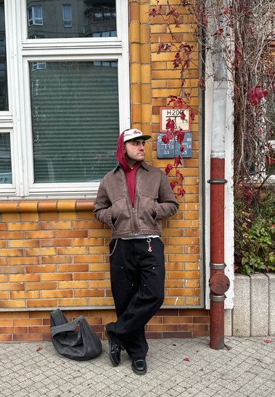 Chaqueta marrón con cremallera sobre un sudadera roja, pantalones anchos negros y zapatos negros; de pie junto a una pared de ladrillo con hojas coloridas y signos.