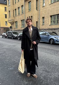 Czarny płaszcz trench na beżowym swetrze dzianinowym, czarne spodnie i brązowe sneakersy. Trzyma jasnobeżową torbę na ramię. Miejski krajobraz w tle.
