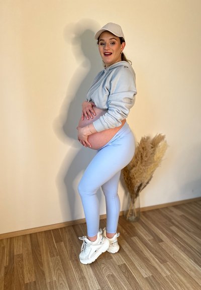 Leggings azul claro combinados con una sudadera gris y zapatillas blancas. Una figura embarazada abraza su barriga, con un fondo claro y una planta decorativa.