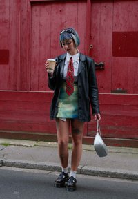 Blazer oversize en cuir noir, chemise blanche, cravate à carreaux rouges, jupe mini dégradé vert-bleu, chaussures compensées noires, sac à main argenté, tenant un café.
