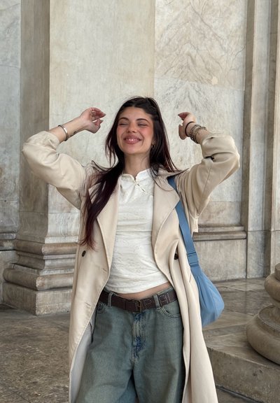 Joven mujer con cabello oscuro sonríe con los ojos cerrados, lleva un abrigo beige, camiseta blanca, vaqueros azules y lleva un bolso azul al hombro, de pie junto a columnas de piedra.