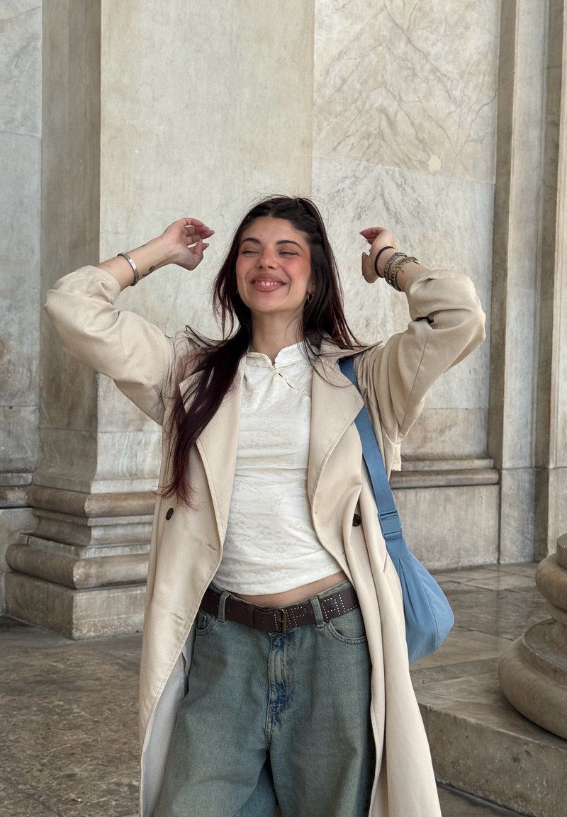 Jeune femme aux cheveux foncés sourit les yeux fermés, portant un manteau beige, un haut blanc, un jean bleu et un sac à épaule bleu, debout près de colonnes en pierre.