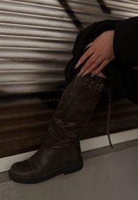 Térdig érő barna bőrcsizma enyhén kopottas hatással, övvel és fém díszítéssel, kerek orral.