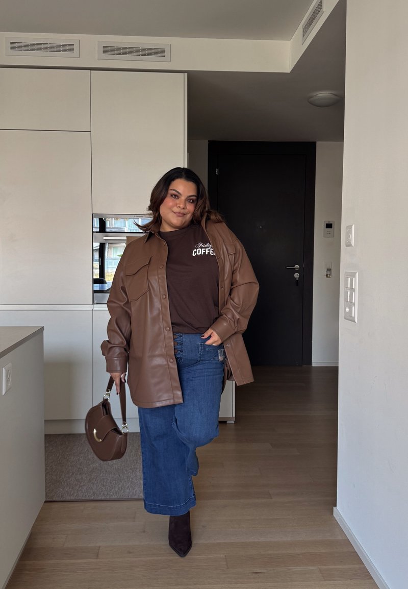 Veste chemise oversize en cuir marron, t-shirt marron foncé avec le texte "café", jeans large en denim bleu, sac à main marron et talons aiguilles foncés.