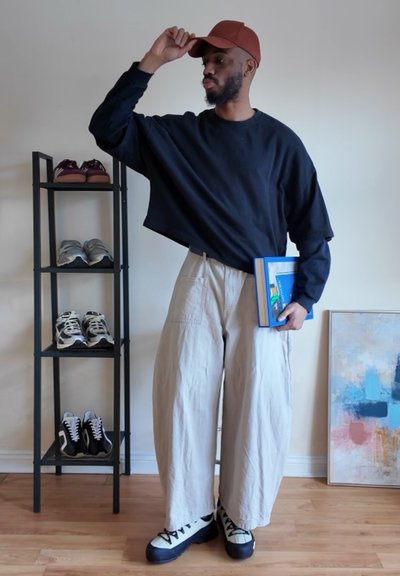 Hombre con gorra roja, camisa negra de manga larga y pantalones anchos beige, sosteniendo libros, de pie junto a un zapatero y una pintura abstracta.