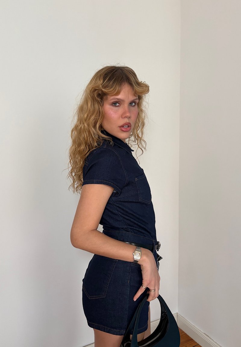 Löysä tummansinisestä denimin materiaalista valmistettu haalari, jossa on lyhyet hihat, mukautuva muotoilu, etutasku ja vyötäröllä oleva vyö. Rakenteellinen ja tekstuuriin perustuva ilme, joka on yhdistetty mustaan käsilaukkuun.