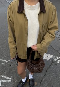 Jakna s olivno zelenim manšetami iz žameta, preko kremastega puloverja, črne kratke hlače in bele nogavice. Drži rjavo usnjeno torbico. Črni mokasini.