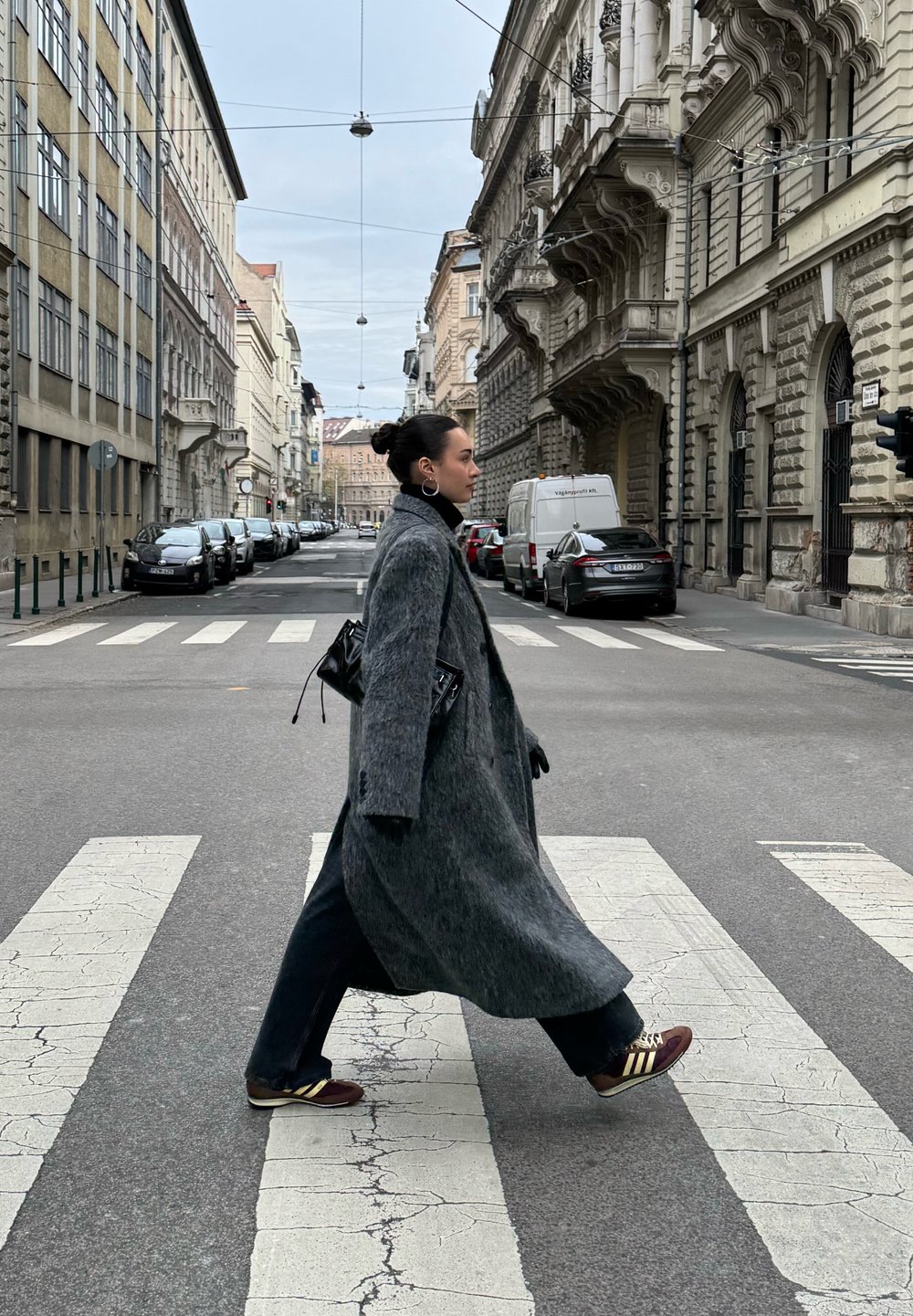 Manteau long gris en tissu texturé, sac noir, pantalon foncé et chaussures marron avec des rayures jaunes, marchant sur un passage pour piétons en milieu urbain.