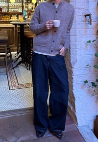 Cárdigan de lana marrón claro con cierre de botones, usado sobre una camisa blanca. Combina con jeans de pierna ancha azul oscuro y zapatos negros. Sostiene una taza.