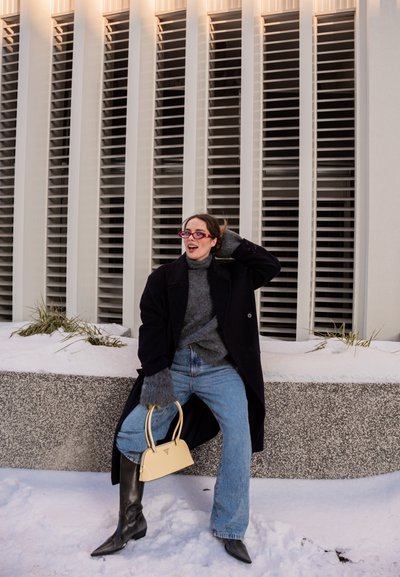 Mujer con abrigo negro, suéter gris, jeans azules y botas negras, sosteniendo un bolso beige, sentada en un borde nevado frente a un edificio con rejillas verticales.