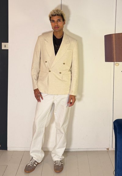 Joven con cabello rizado rubio que lleva un blazer cruzado beige, pantalones blancos y zapatillas grises, de pie en interiores.