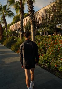 Črna lahka jakna, črne kratke hlače in bele nogavice. Pot obdana z živahnimi cvetlicami in palmami v svetli zunanji nastavitvi.