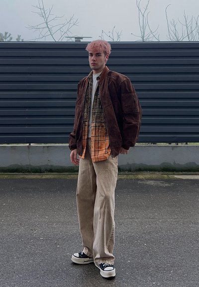 Persona joven con cabello rosa de pie al aire libre, vestida con chaqueta marrón, camisa de cuadros naranja, pantalones anchos beige y zapatillas blancas y negras.