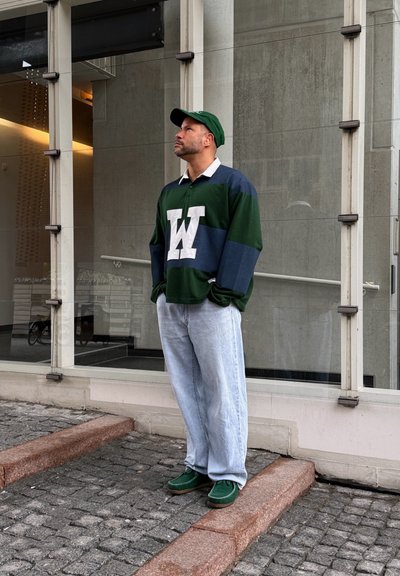 Hombre con camiseta de rugby a rayas verdes y azul marino con una gran "W" blanca, gorra verde, vaqueros azul claro y zapatos verdes, de pie sobre adoquines cerca de un edificio de cristal.
