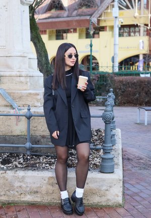 Mujer con blazer negro, vestido, medias y gafas de sol, de pie junto a una jardinera de piedra sosteniendo una taza de café en una calle de la ciudad.