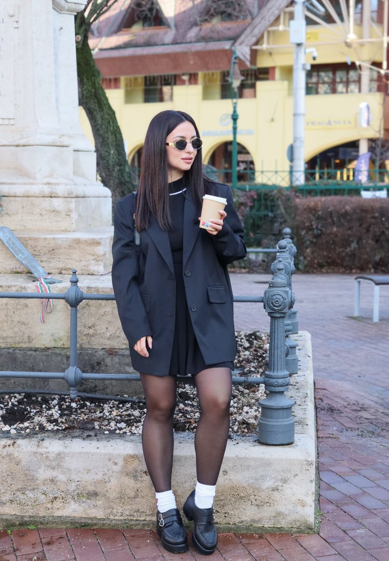 Woman wearing black blazer, dress, stockings, and sunglasses stands near stone planter holding a coffee cup on a city street.
