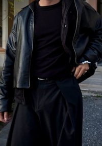 Persona che indossa una giacca di pelle nera, una maglietta nera e pantaloni larghi neri, in piedi all'aperto vicino a gradini di cemento.