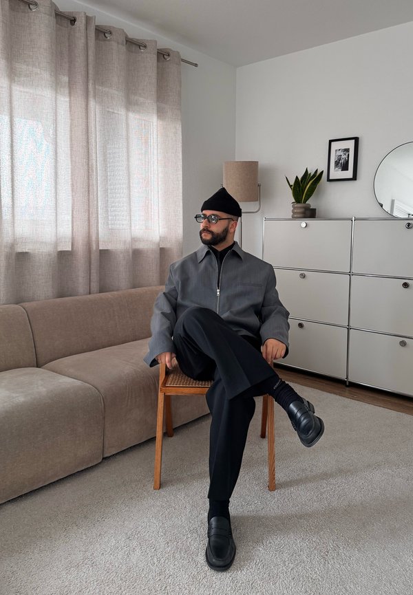 Egy szemüveges férfi, szürke dzsekiben, fekete nadrágban és kalapban keresztbe tett lábakkal ül egy fa széken egy modern nappaliban, kanapéval és szekrénnyel.