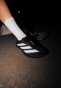 Černá sportovní obuv s bílými pruhy, textilním svrškem a texturovanou podrážkou se vzorem kruhů, nošená s bílou ponožkou na tmavém povrchu.