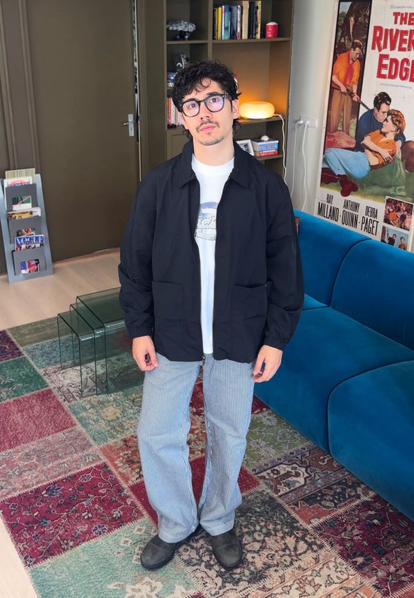 Junger Mann mit Brille, trägt eine schwarze Jacke, ein weißes Hemd und gestreifte Hosen, steht in einem Wohnzimmer mit buntem Teppich.