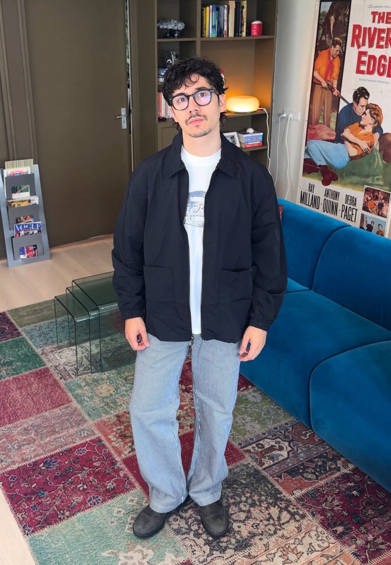 Jeune homme avec des lunettes portant une veste noire, une chemise blanche et un pantalon rayé, debout dans un salon recouvert d'un tapis coloré.