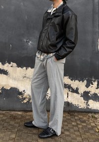 Άνδρας με μαύρο δερμάτινο μπουφάν, πουκάμισο καρό, φαρδιά γκρι παντελόνια και μαύρα παπούτσια με αγκράφα στέκεται μπροστά σε φθαρμένο ασπρόμαυρο τοίχο.