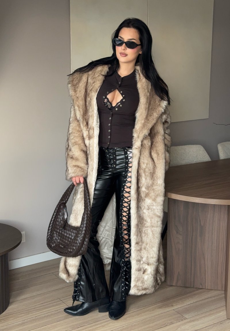 Femme portant un long manteau de fourrure beige, pantalon en cuir noir lacé, haut noir avec découpe, lunettes de soleil, et tenant un sac à main tressé marron à l'intérieur.