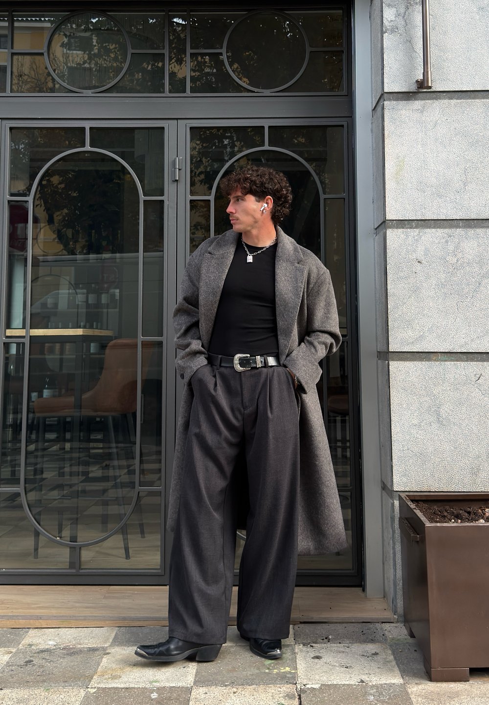 Lang grå kåpe over en svart skjorte, kombinert med mørke vide bukser. Tilbehør inkluderer belte, kjede og ørepropper. Står nær en glassdør.