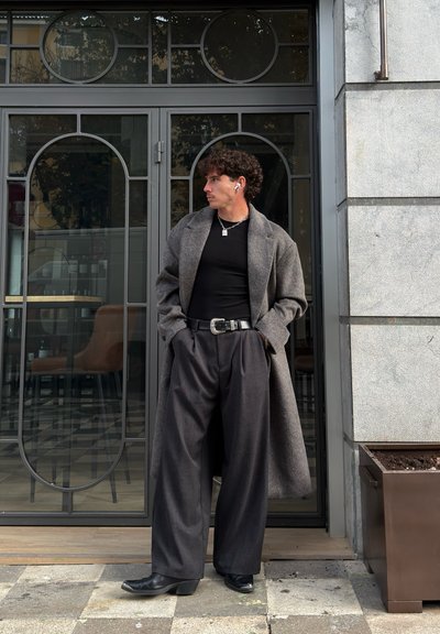 Abrigo largo gris sobre una camisa negra, combinado con pantalones anchos oscuros. Accesorios incluyen un cinturón, una cadena y auriculares. De pie cerca de una puerta de vidrio.