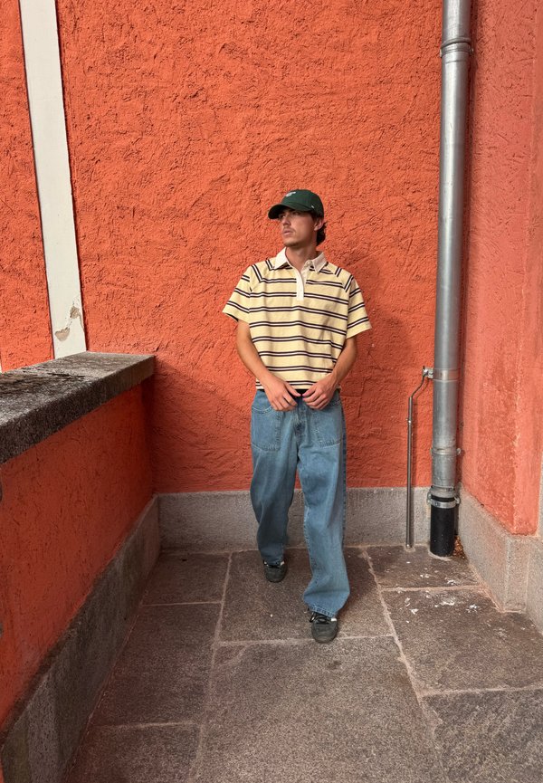 Jovem vestindo boné verde, camisa amarela às riscas e jeans azuis largos a caminhar junto a uma parede laranja texturada e um tubo metálico num piso de pedra.
