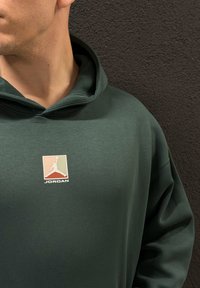Grønn hettegenser laget av mykt materiale med en logo-patch som viser en silhuett og teksten "JORDAN" i hvitt, plassert mot en teksturert mørk bakgrunn.