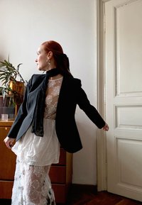 Sort blazer over en hvit blonderbluse med blomsterpattern, lagvis hvit skjørt. Svart skjerf fremhever utseendet. Varm innendørs belysning.
