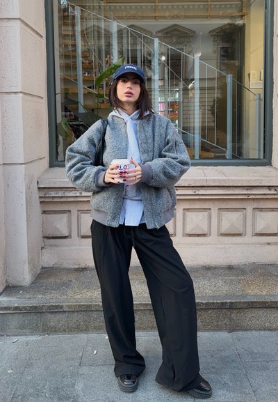 Chaqueta bomber gris sobre una sudadera ligera gris, pantalones anchos negros y una gorra azul marino. Sosteniendo una taza impresa, de pie frente a un edificio.