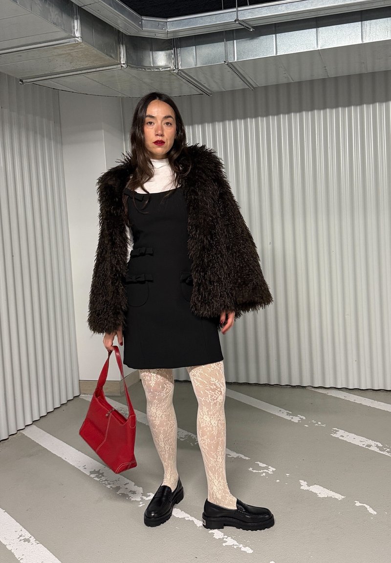 Frau steht in einer Tiefgarage, trägt ein schwarzes Kleid, braunen Pelzmantel, weiße Spitzenstrumpfhosen, schwarze Schuhe und hält eine rote Handtasche.