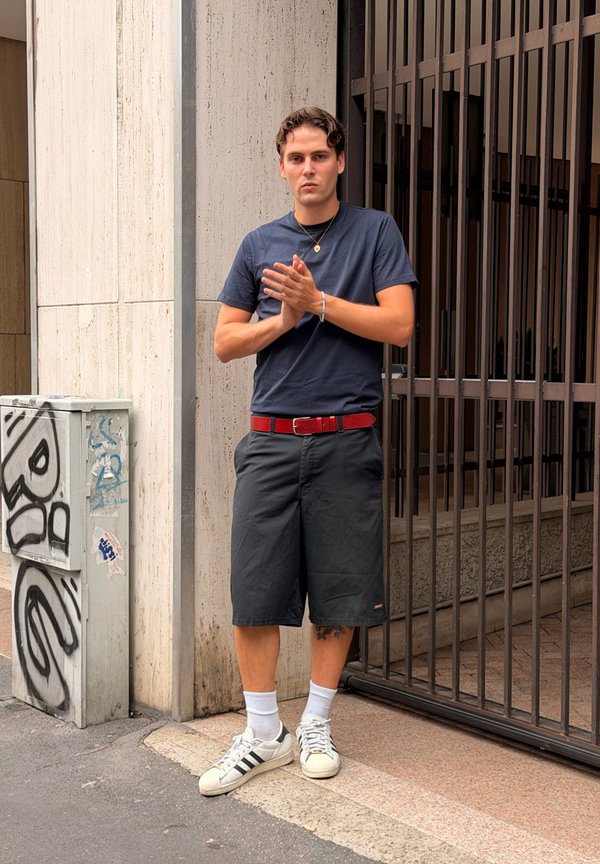Ung mand, der klapper hænder, iført navy skjorte, sorte shorts, hvide sneakers, står ved en metalport og et gadeskab dækket med graffiti.