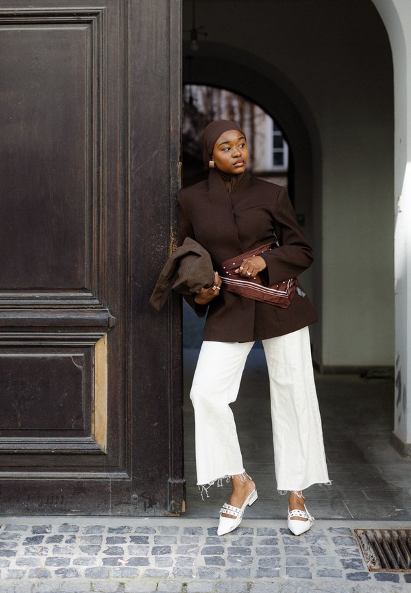 Donna con hijab marrone e giacca, pantaloni bianchi a gamba larga e scarpe bianche con borchie appoggiata a una grande porta di legno in un ingresso ad arco.