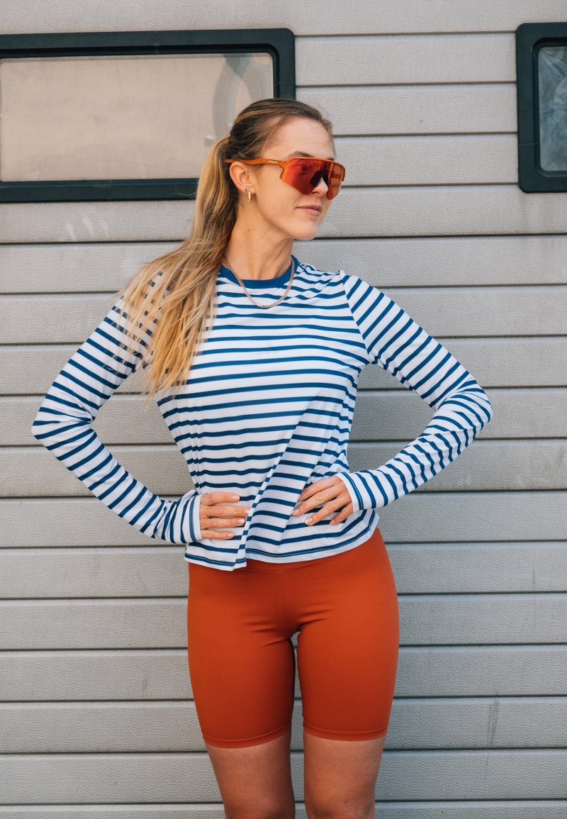 Femme aux longs cheveux blonds attachés en queue de cheval, portant des lunettes de soleil orange, un t-shirt à manches longues rayé bleu et blanc, et un short de cyclisme orange, se tient les mains sur les hanches.