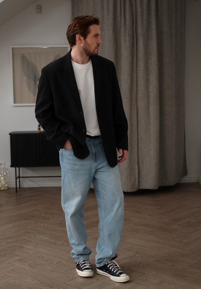 Blazer negro sobre camiseta blanca, jeans de mezclilla azul claro y zapatillas negras. Habitación con suelo de madera y cortinas grises de fondo.