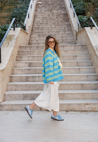 Cárdigan de punto a rayas azul y amarilla, pantalones de pierna ancha blancos y zapatillas azules con cordones, bajando unos escalones de piedra rodeados de vegetación.