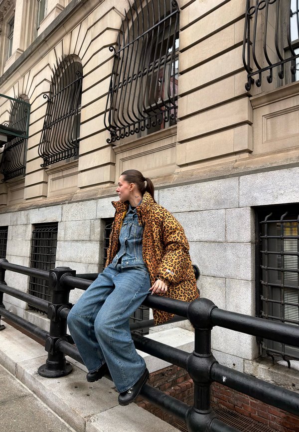 Young woman with ponytail wearing leopard print jacket and denim outfit sitting on black metal railing in front of stone building with barred windows.