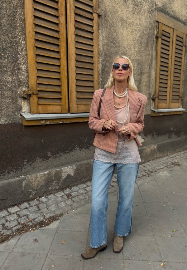 Femme blonde portant des lunettes de soleil, des colliers superposés, un blazer rose à rayures, un top en dentelle, un jean large et des bottes marron, debout dans une rue pavée.