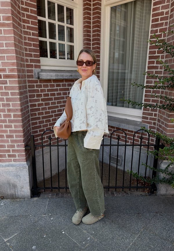 Femme portant des lunettes de soleil, un chemisier blanc texturé, un pantalon large vert et des chaussures beiges, debout à l'extérieur près d'un mur en briques et d'une fenêtre, tenant un sac à main marron.