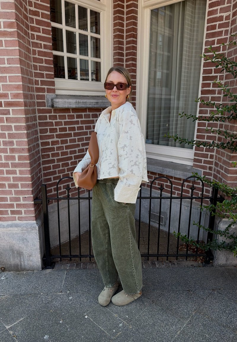 Femme portant des lunettes de soleil, un chemisier blanc texturé, un pantalon large vert et des chaussures beiges, debout à l'extérieur près d'un mur de briques et d'une fenêtre, tenant un sac à main marron.